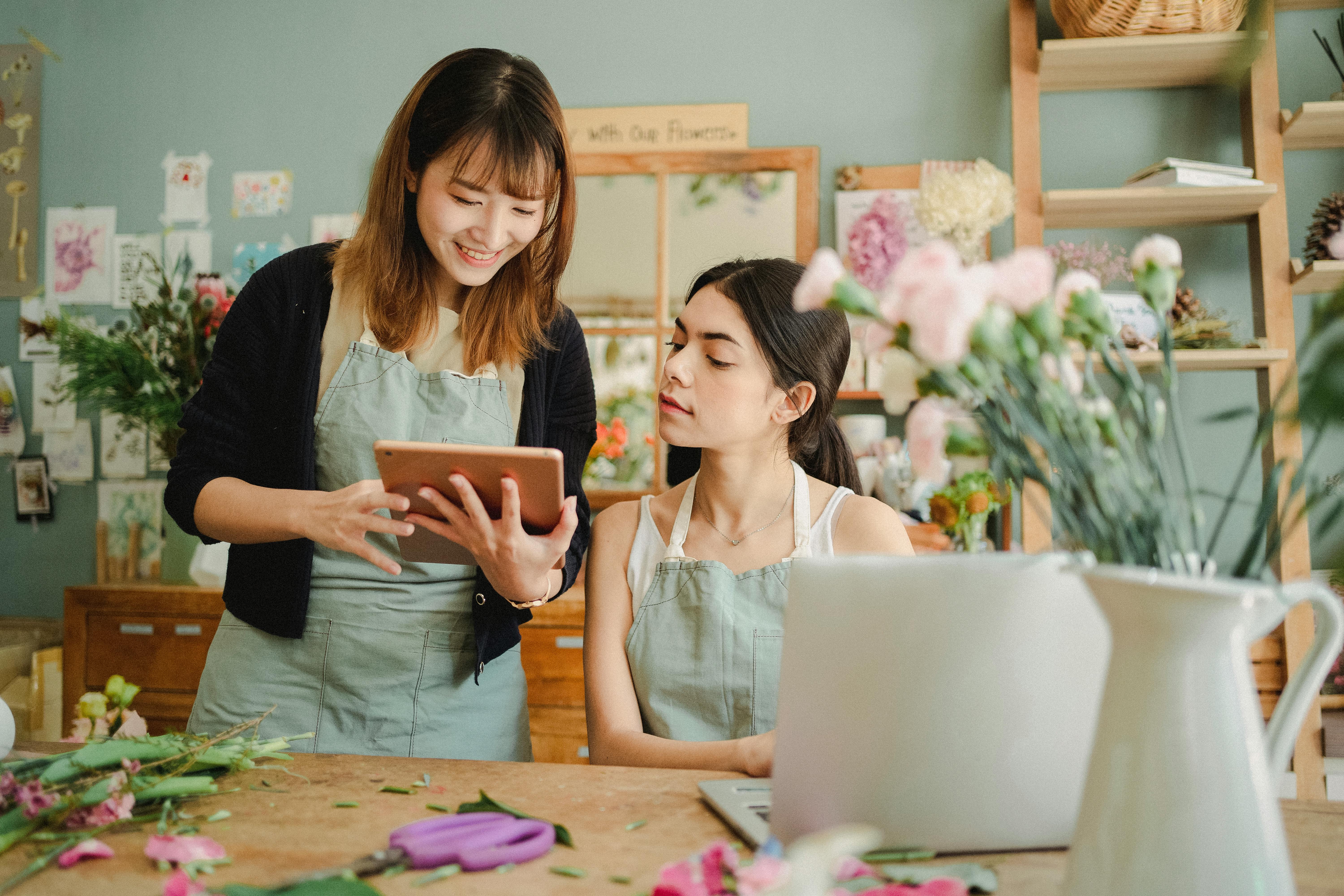 Content multiethnic female coworkers at table with flowers surfing tablet while choosing design for bouquet during work in florist shop