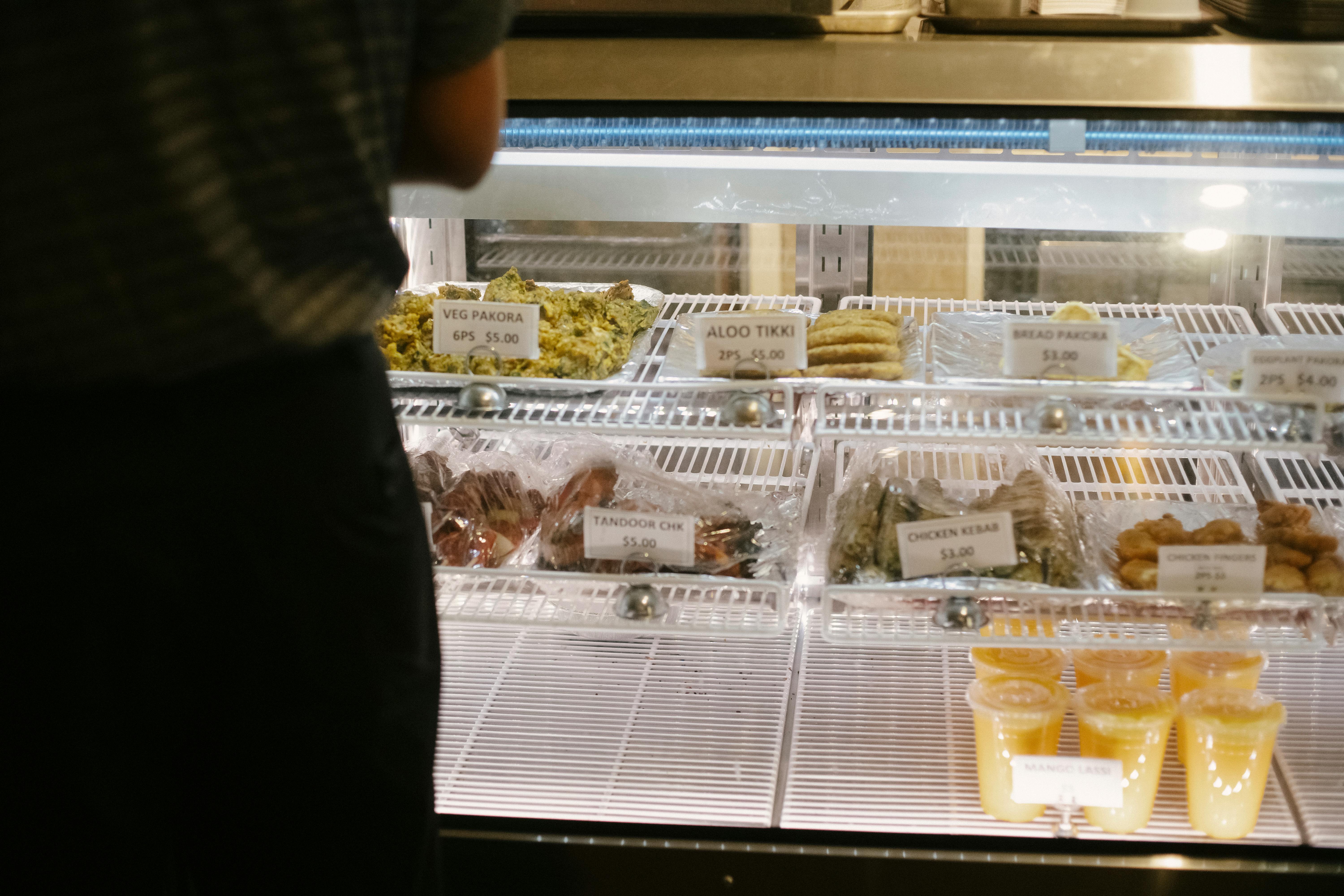 Back view of anonymous customer standing near glass showcase with assorted sweets while choosing delicious desserts in confectionery shop and making order