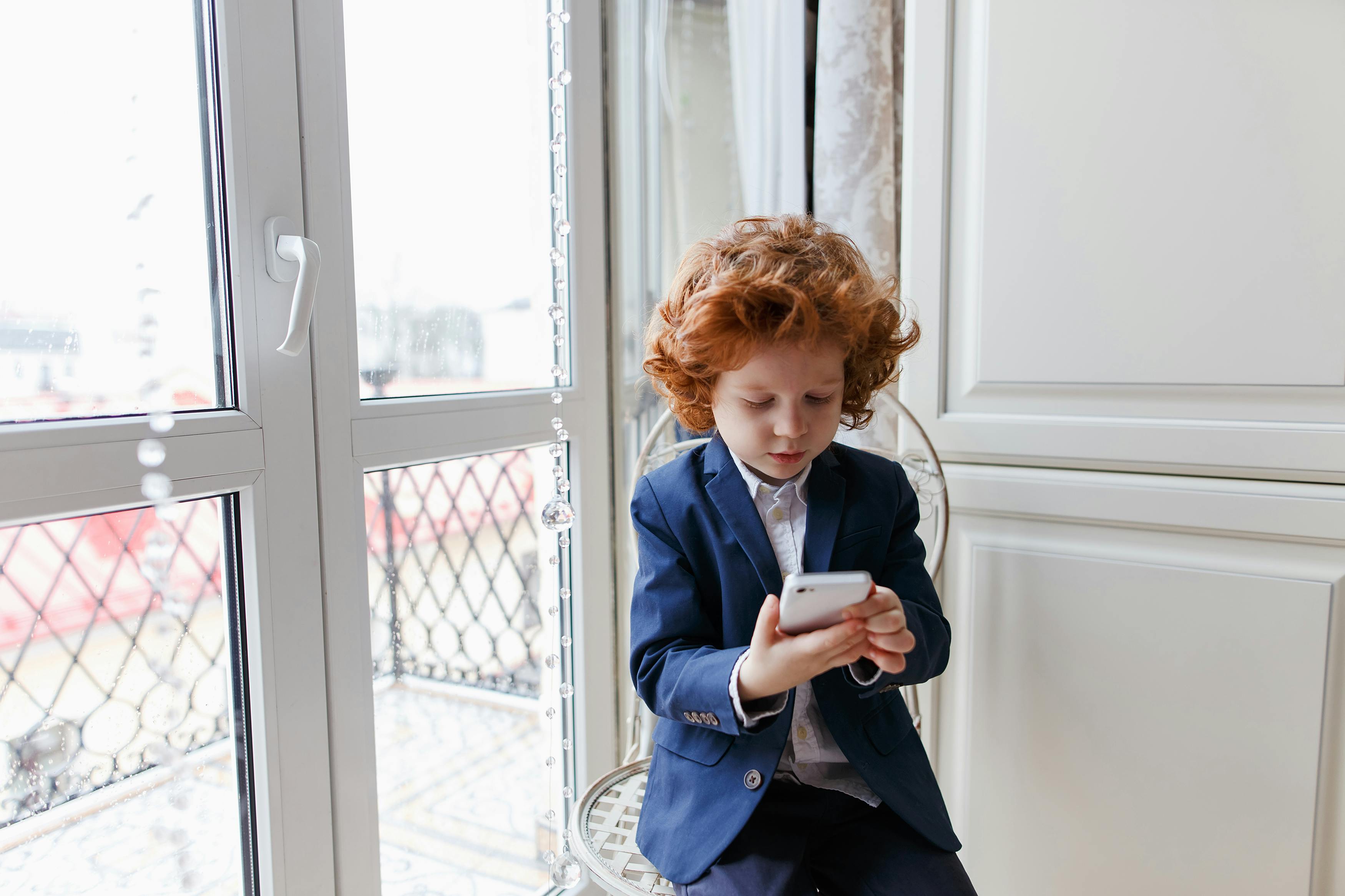 Little redhead boy uses a smartphone sitting in his room at home