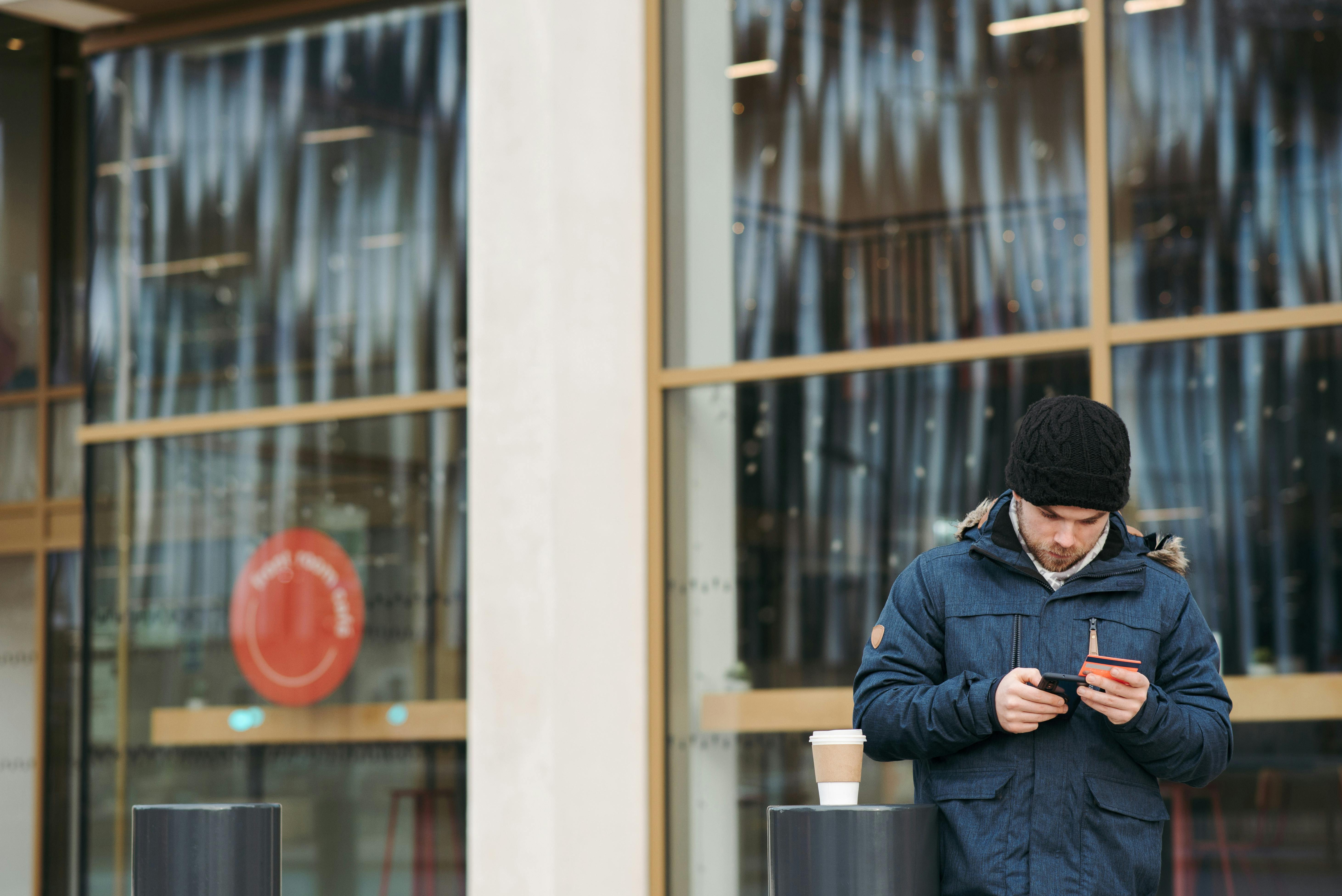 Focused young male in warm outerwear and hat standing near modern building with takeaway coffee and using smartphone while making online shopping with credit card